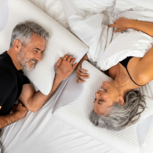 Vue de dessus d'un couple d'une cinquantaine d'années, souriants et détendus, allongés dans un lit blanc. Ils utilisent chacun un oreiller cervical ergonomique OrthoDodo à mémoire de forme pour un soutien optimal de la nuque.