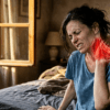 Femme au réveil assise sur son lit souffrant d'un torticolis intense, la main sur la nuque. Une illustration anatomique rouge en surimpression montre les muscles cervicaux contractés. Lumière matinale dorée et ambiance cinématographique.