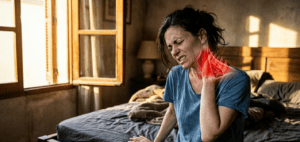 Femme au réveil assise sur son lit souffrant d'un torticolis intense, la main sur la nuque. Une illustration anatomique rouge en surimpression montre les muscles cervicaux contractés. Lumière matinale dorée et ambiance cinématographique.